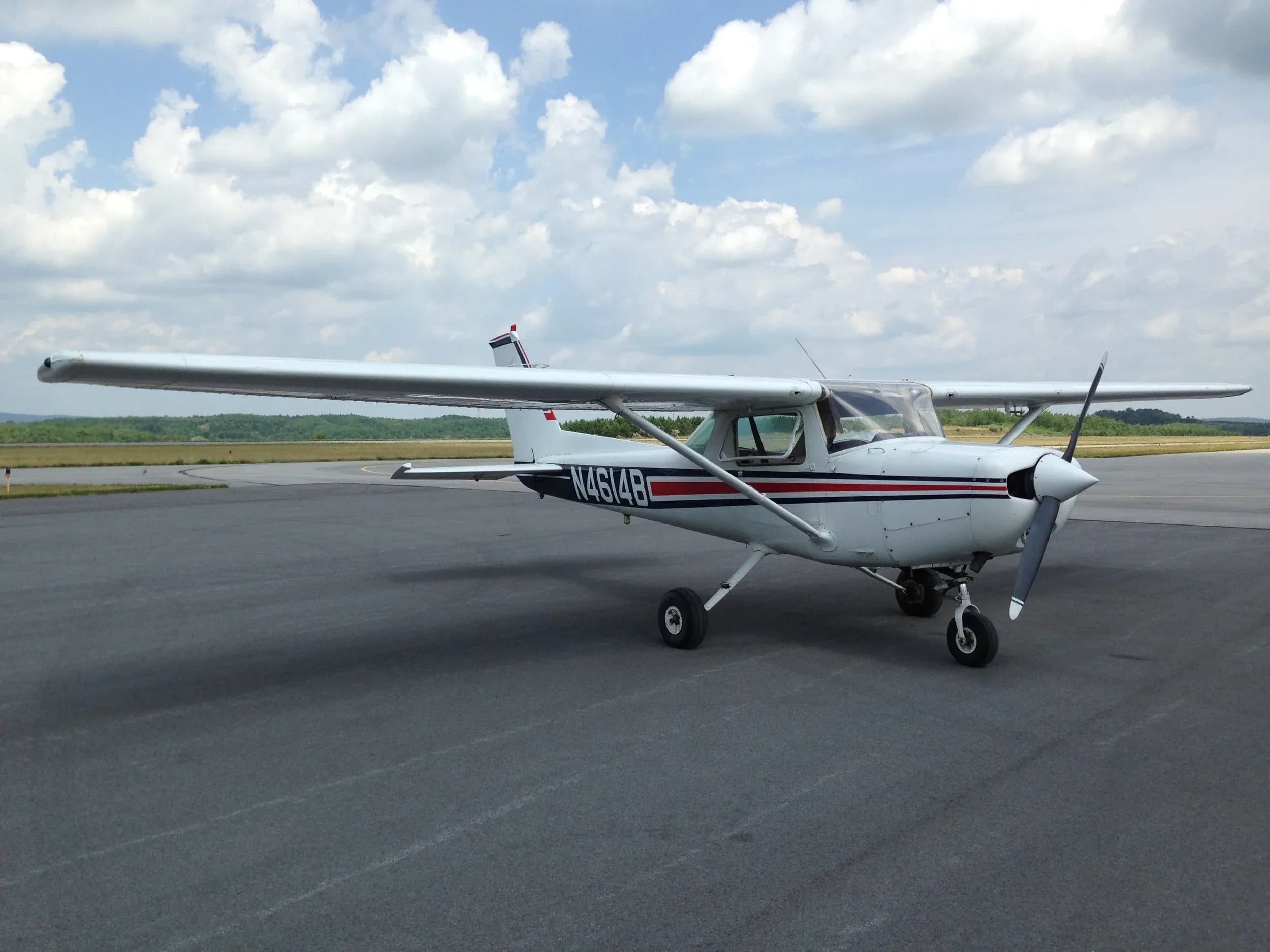A white single-engine Cessna aircraft with red and black stripes on its fuselage is parked on an asphalt tarmac under a partly cloudy sky.