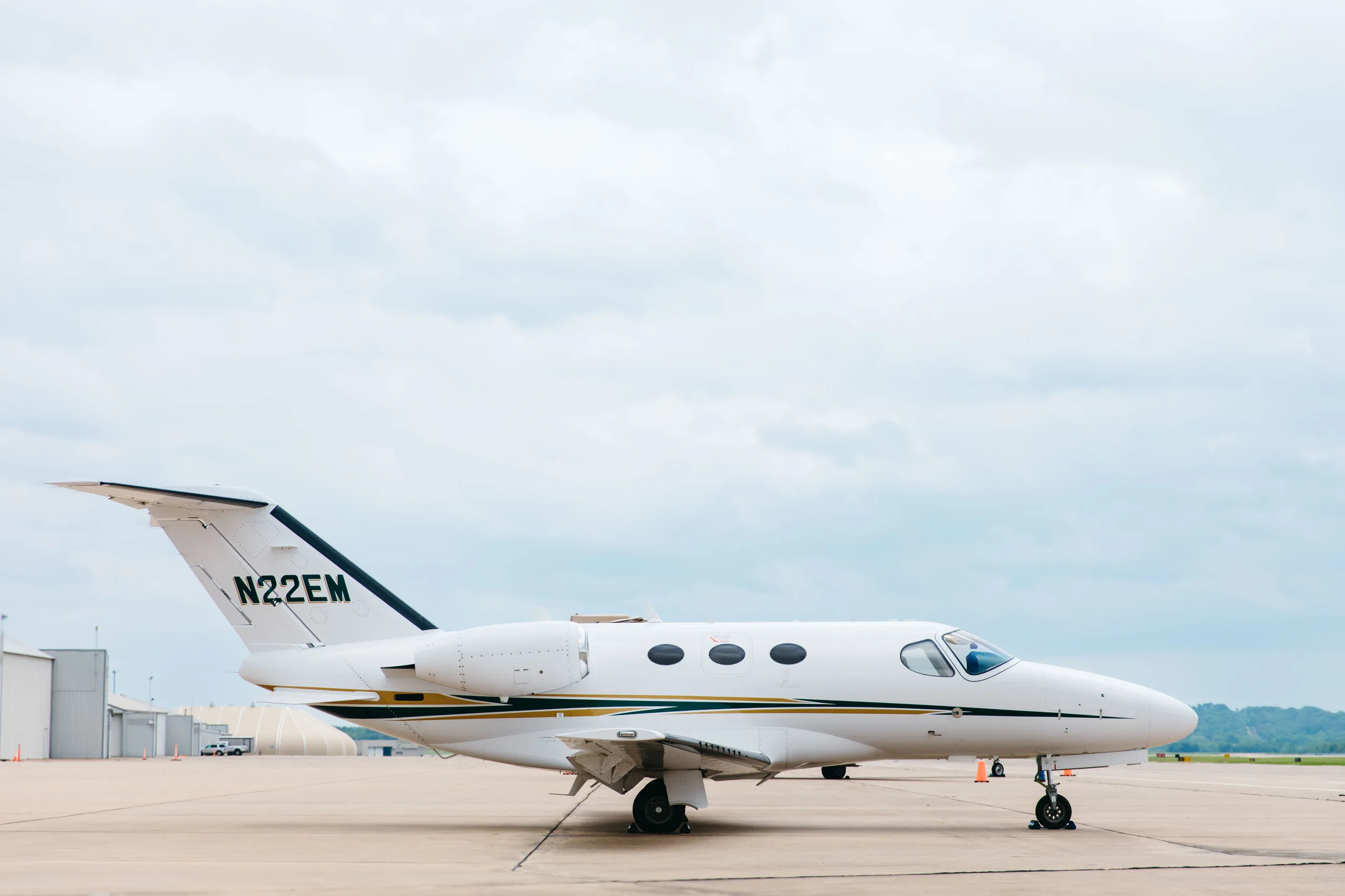A side profile of a white Cessna Citation M2 business jet, tail number N22EM, with gold and dark green stripes, parked on a tarmac with distant hangars.