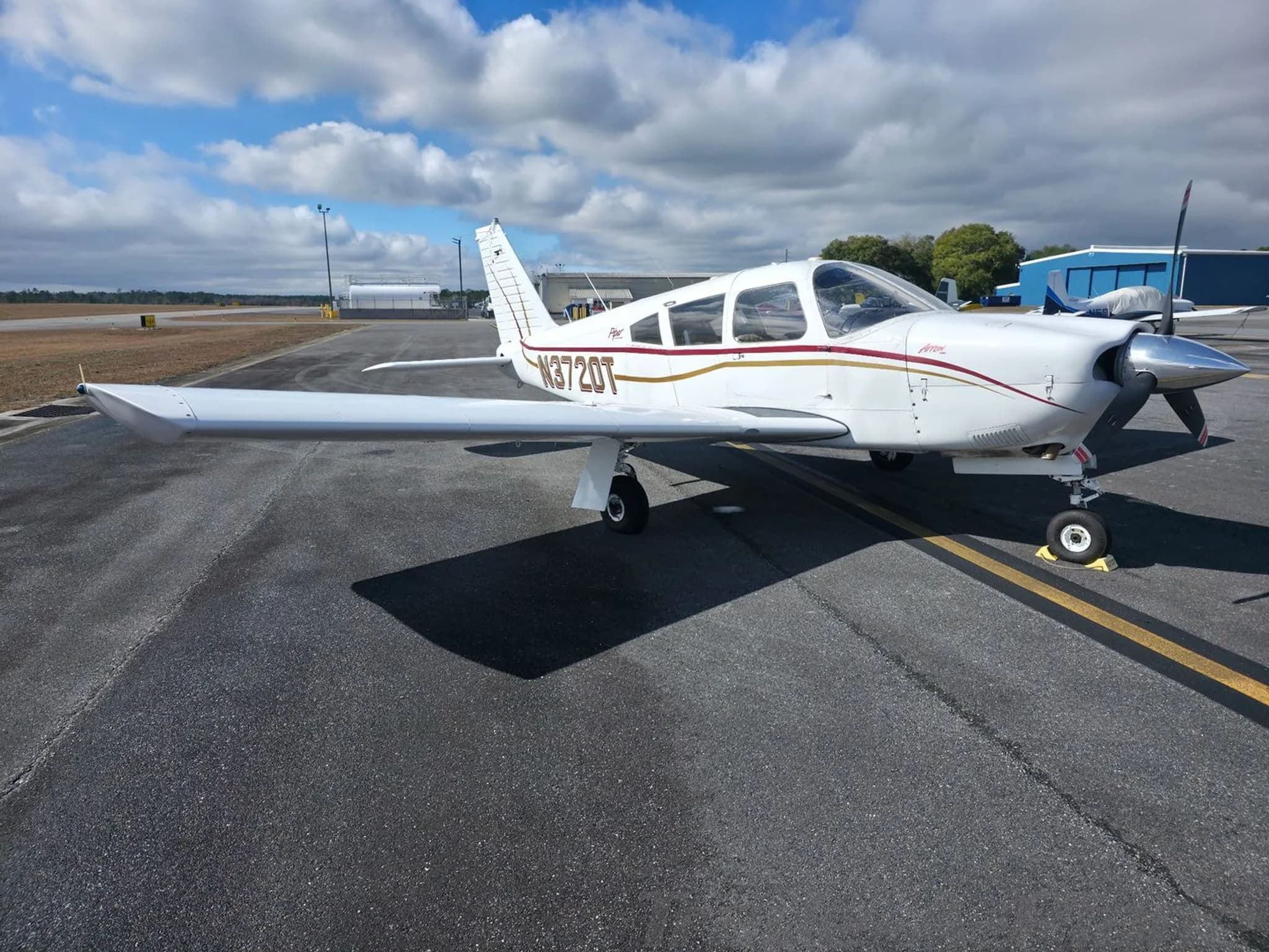 A white Piper Arrow aircraft with gold and red stripes, registration N3720T, parked on an asphalt apron under a partly cloudy sky.