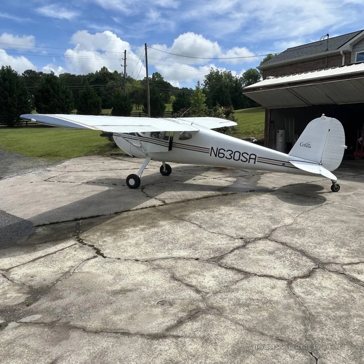 A white Cessna 170B with dark blue and gold stripes is parked on a cracked concrete pad under a partly cloudy sky.