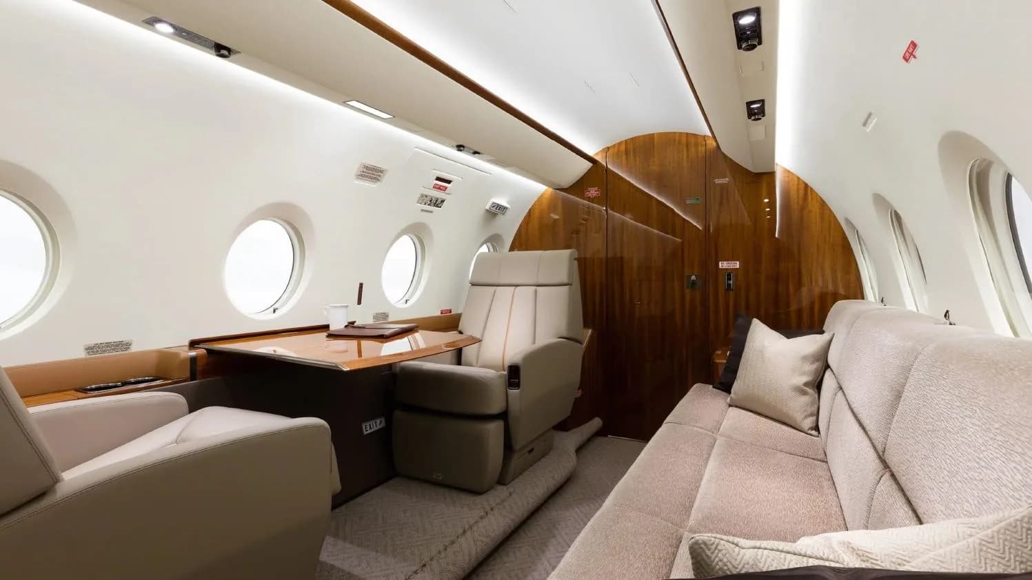 A section of an aircraft cabin featuring a beige sofa, a single seat, and a table with wood paneling.