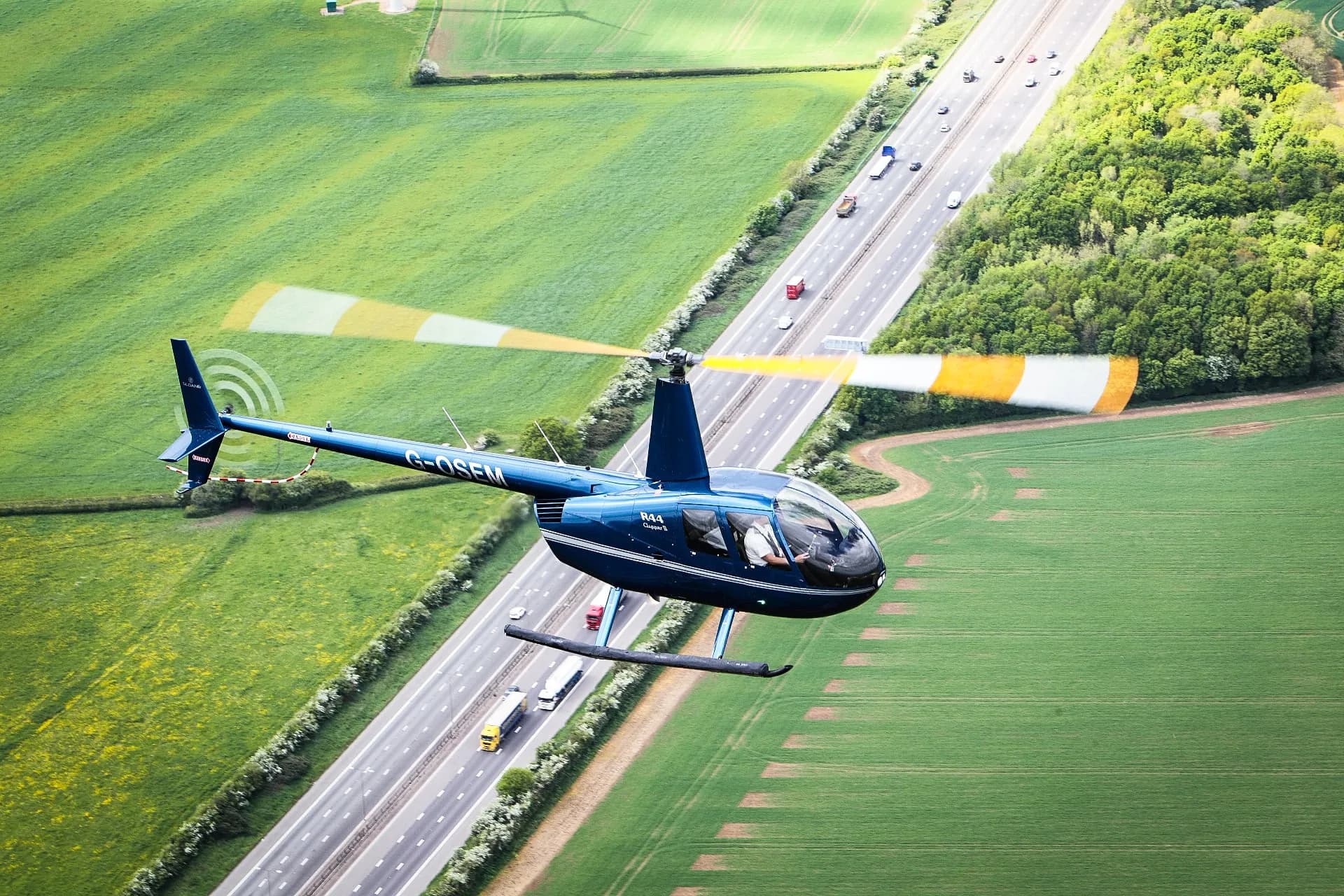 A blue Robinson R44 helicopter flying over a highway and fields.
