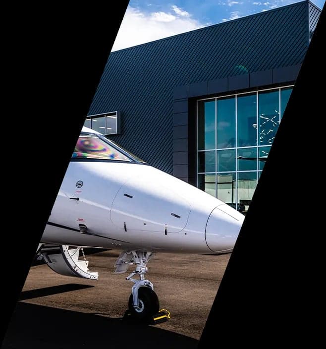 Front view of a white private jet's nose and landing gear, parked in front of a modern hangar.