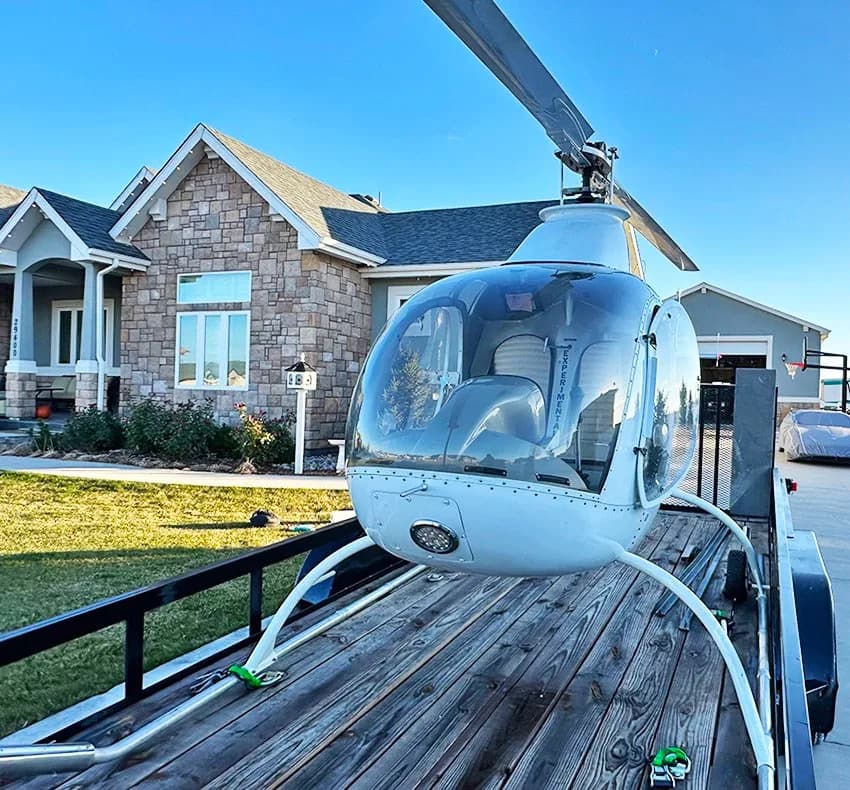Front view of a white helicopter on a wooden trailer, parked in front of a large stone house.