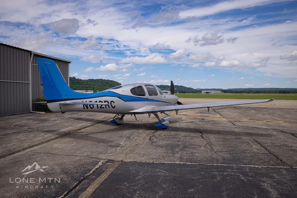 A silver and blue Cirrus SR22T Carbon aircraft, registration N612RC, parked on an airfield next to a hangar.