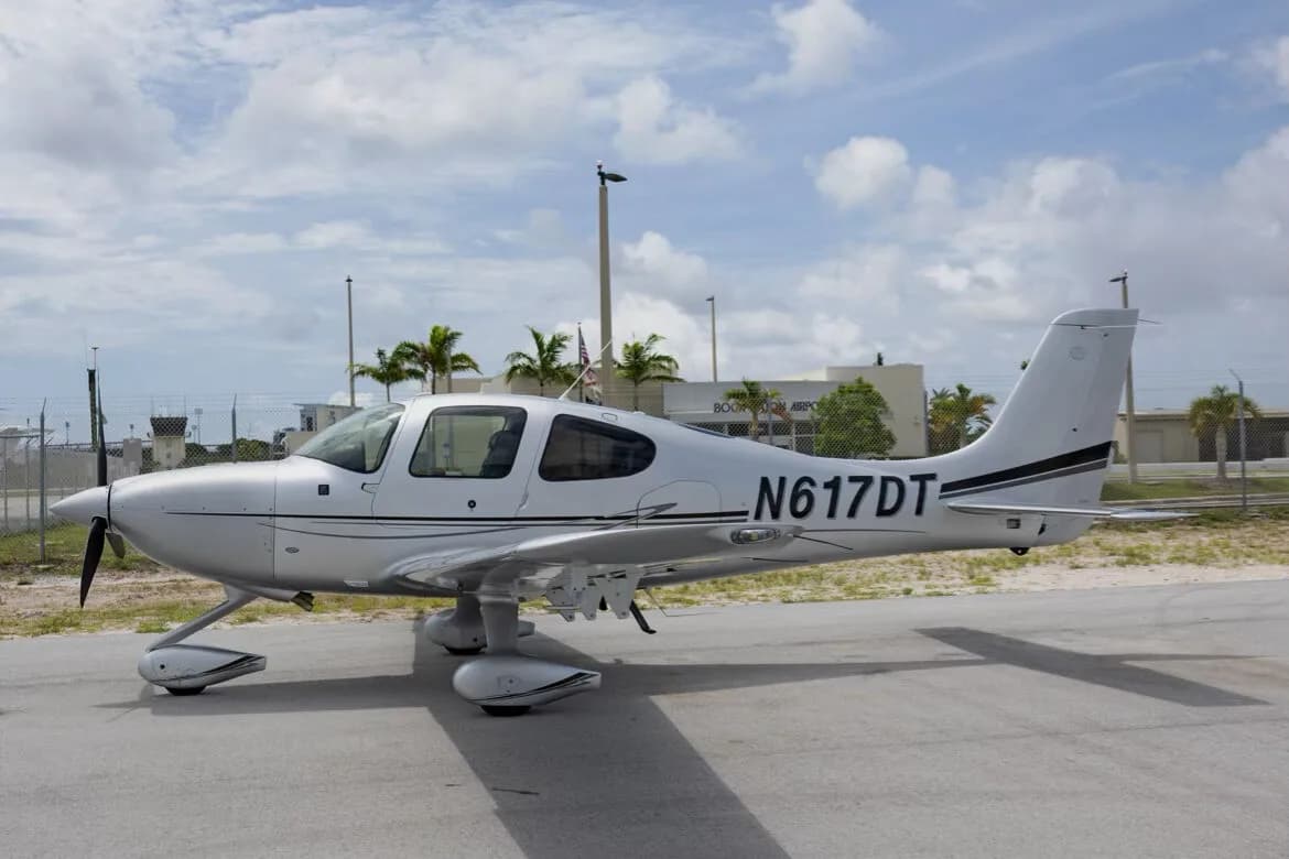 A silver Cirrus SR22 aircraft with tail number N617DT parked on an airport tarmac with palm trees and buildings in the background.