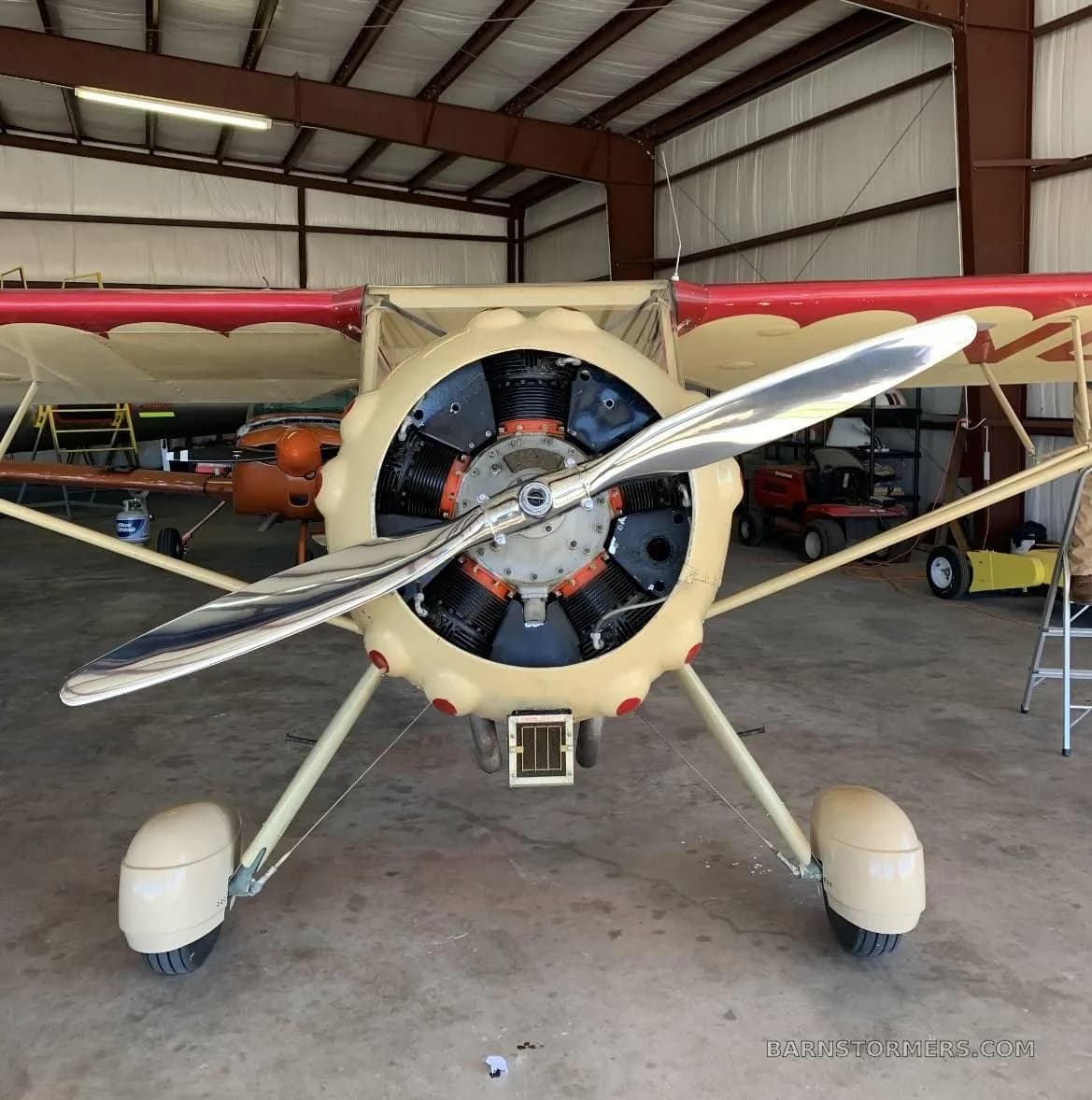 Front view of a cream-colored vintage aircraft with a polished propeller and exposed radial engine, parked inside a hangar.