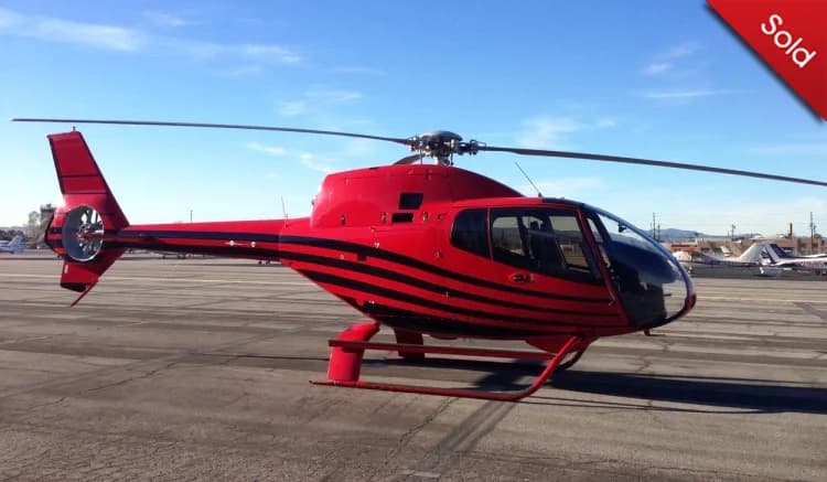 A red helicopter with black horizontal stripes is parked on an asphalt airfield under a clear blue sky.