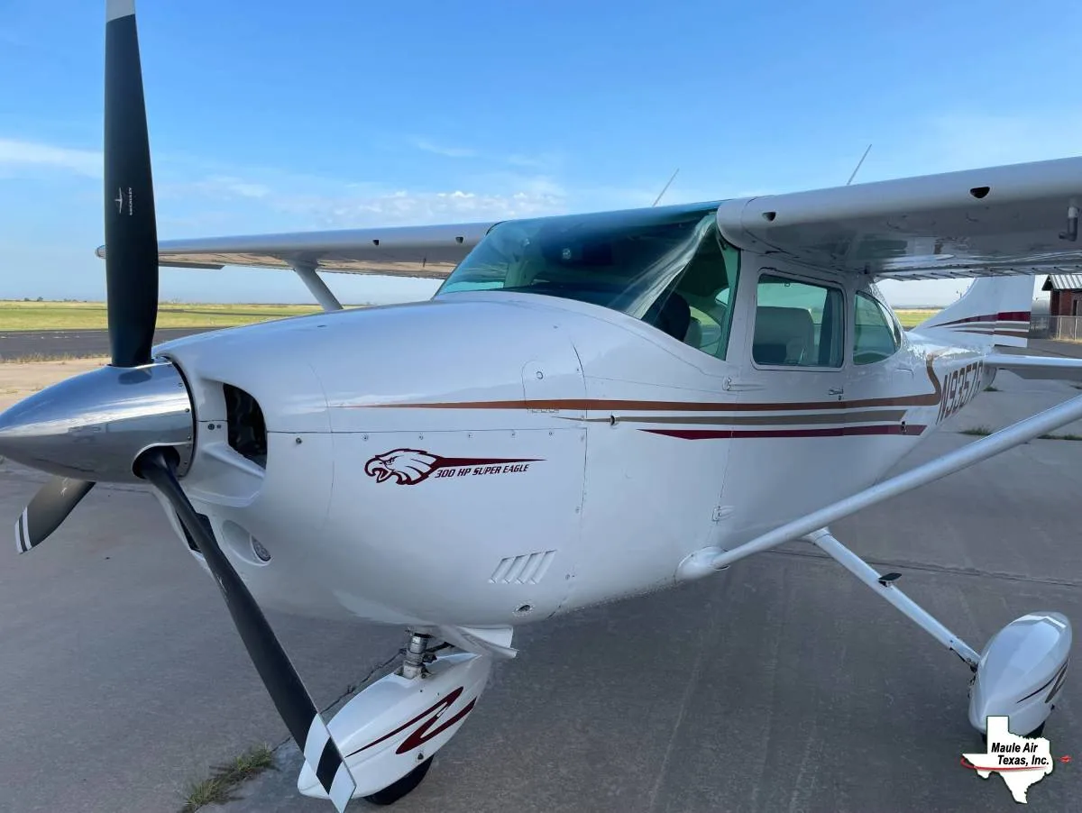 A white Cessna 300 HP Super Eagle with brown and gold stripes is parked on an airfield under a blue sky, viewed from the front-left.