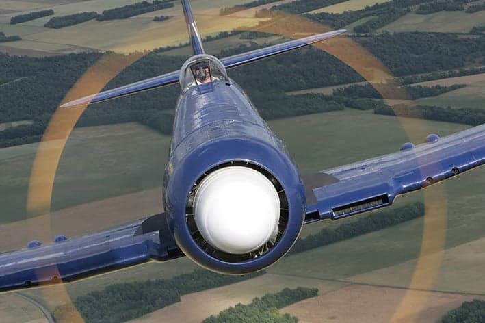 A blue aircraft flying over a rural landscape, leaving a smoke trail.
