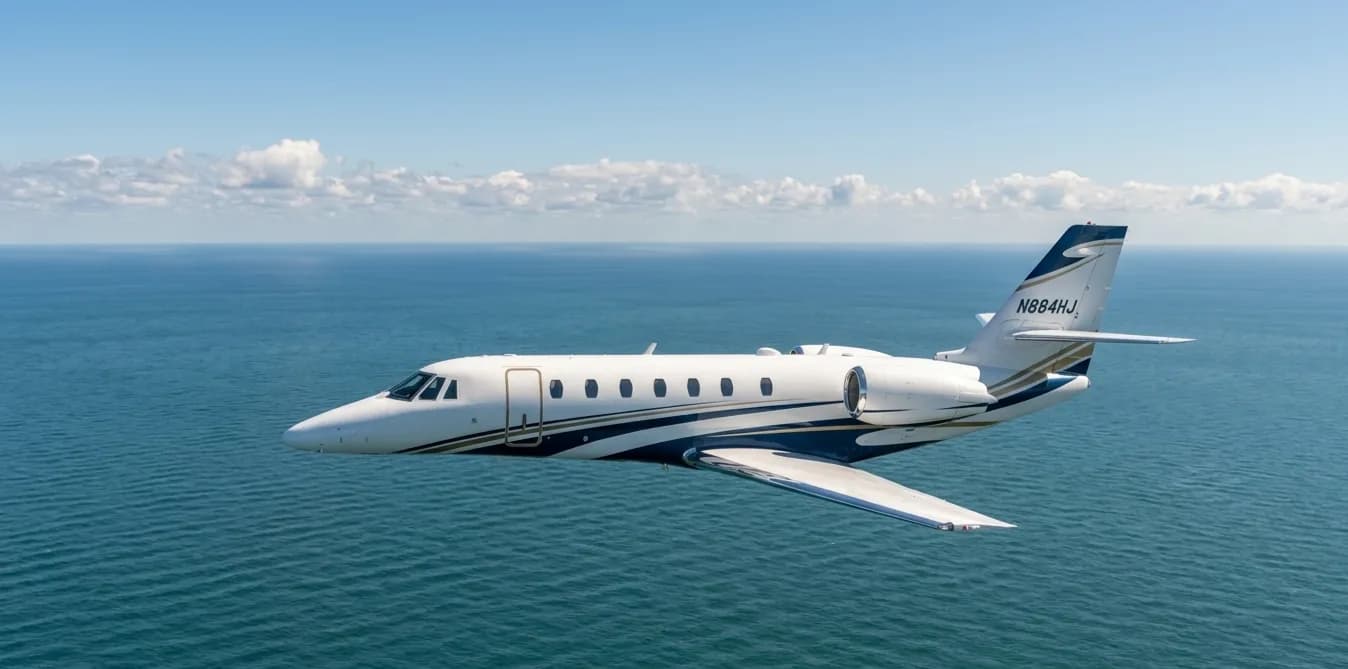 A white business jet with blue and gold stripes, registration N884HJ, flies over a blue ocean under a partly cloudy sky.