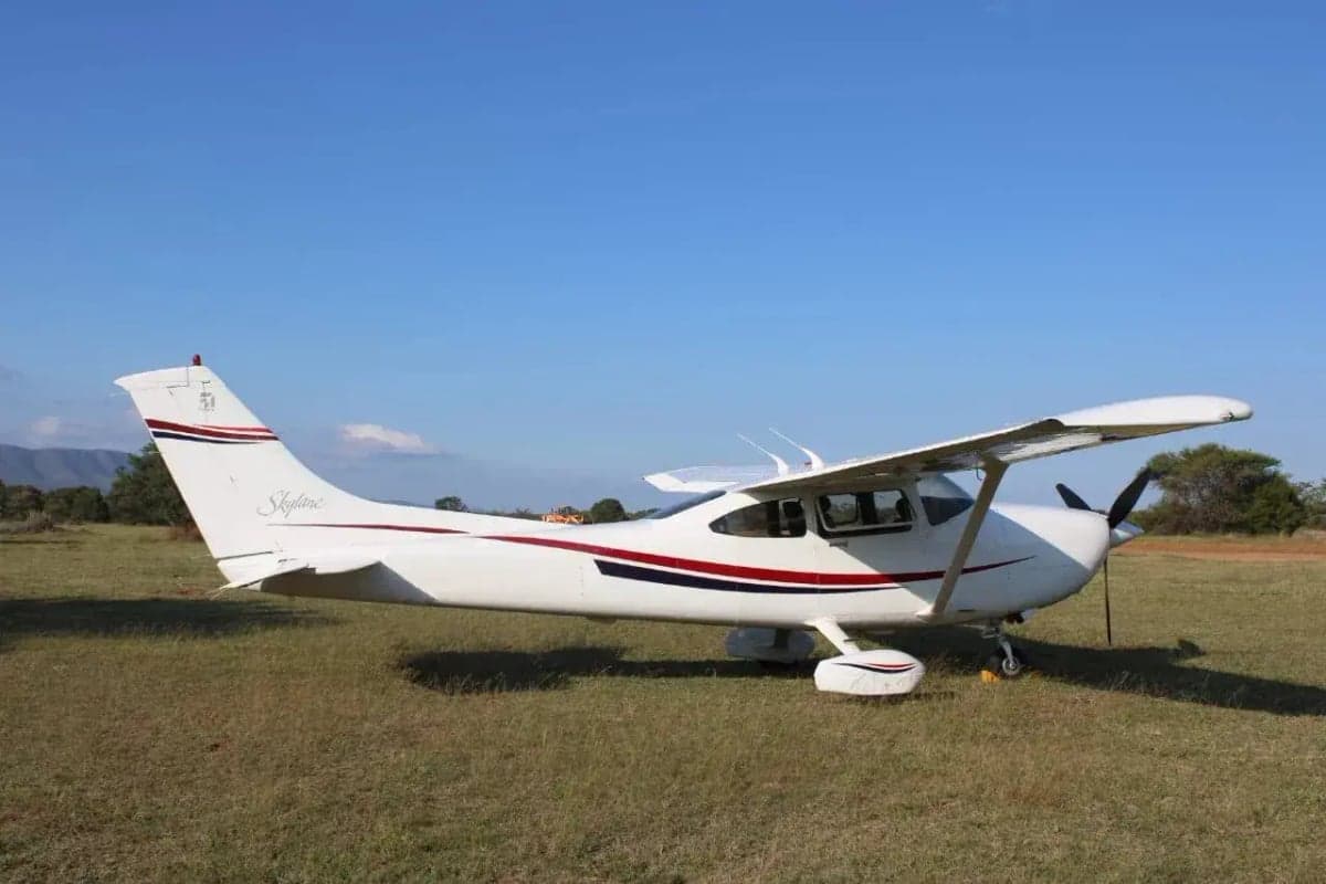 Side view of a Cessna aircraft parked on a grassy field, featuring its white paint scheme with red and blue accents.