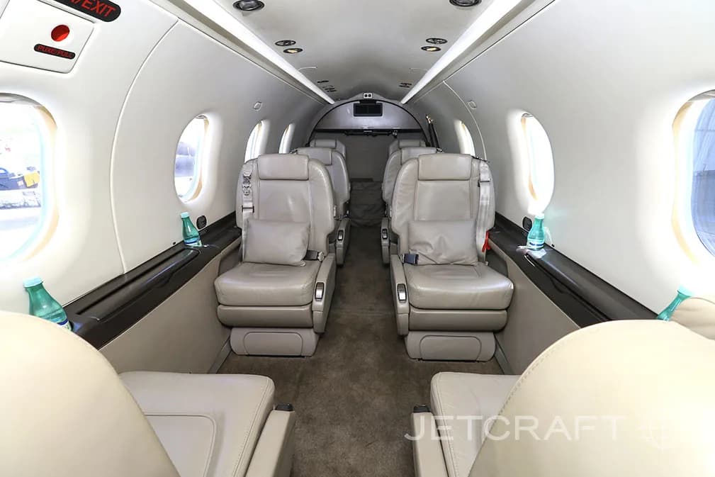 The interior cabin of a private jet features light beige leather seats arranged in a club configuration with small windows.