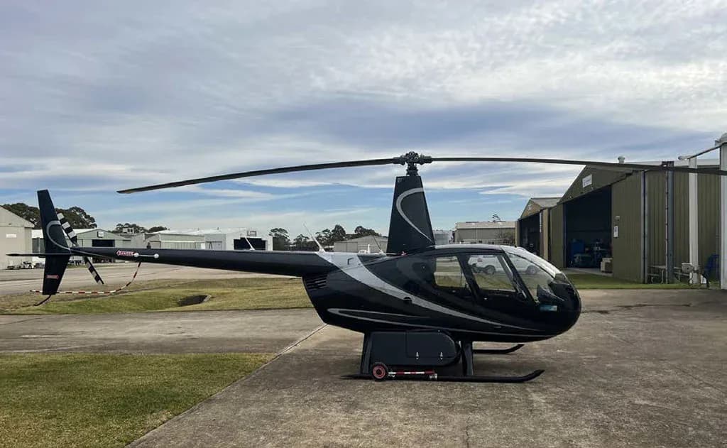 A black Robinson R44 helicopter with silver stripes on a tarmac, viewed from the left side.