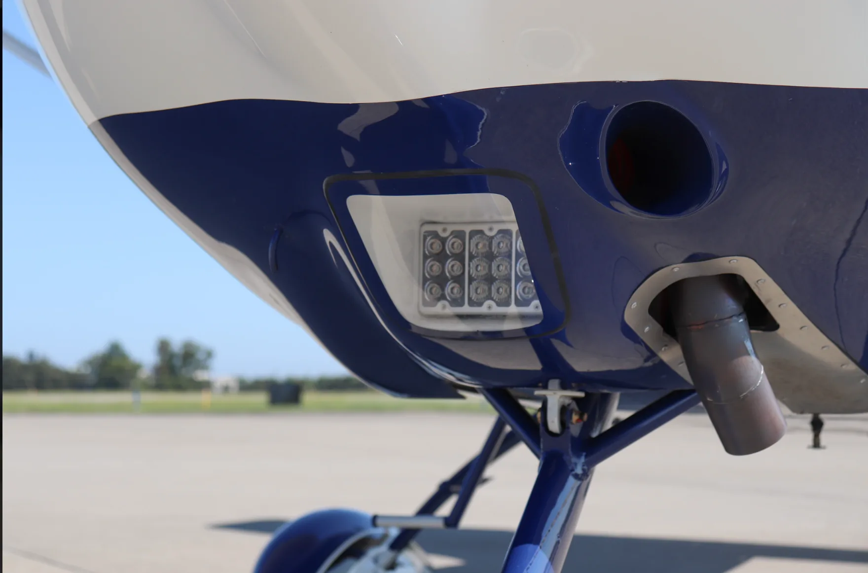 A close-up of the underside of the white and blue Tecnam P2010 fuselage, showing an LED light and exhaust pipe.