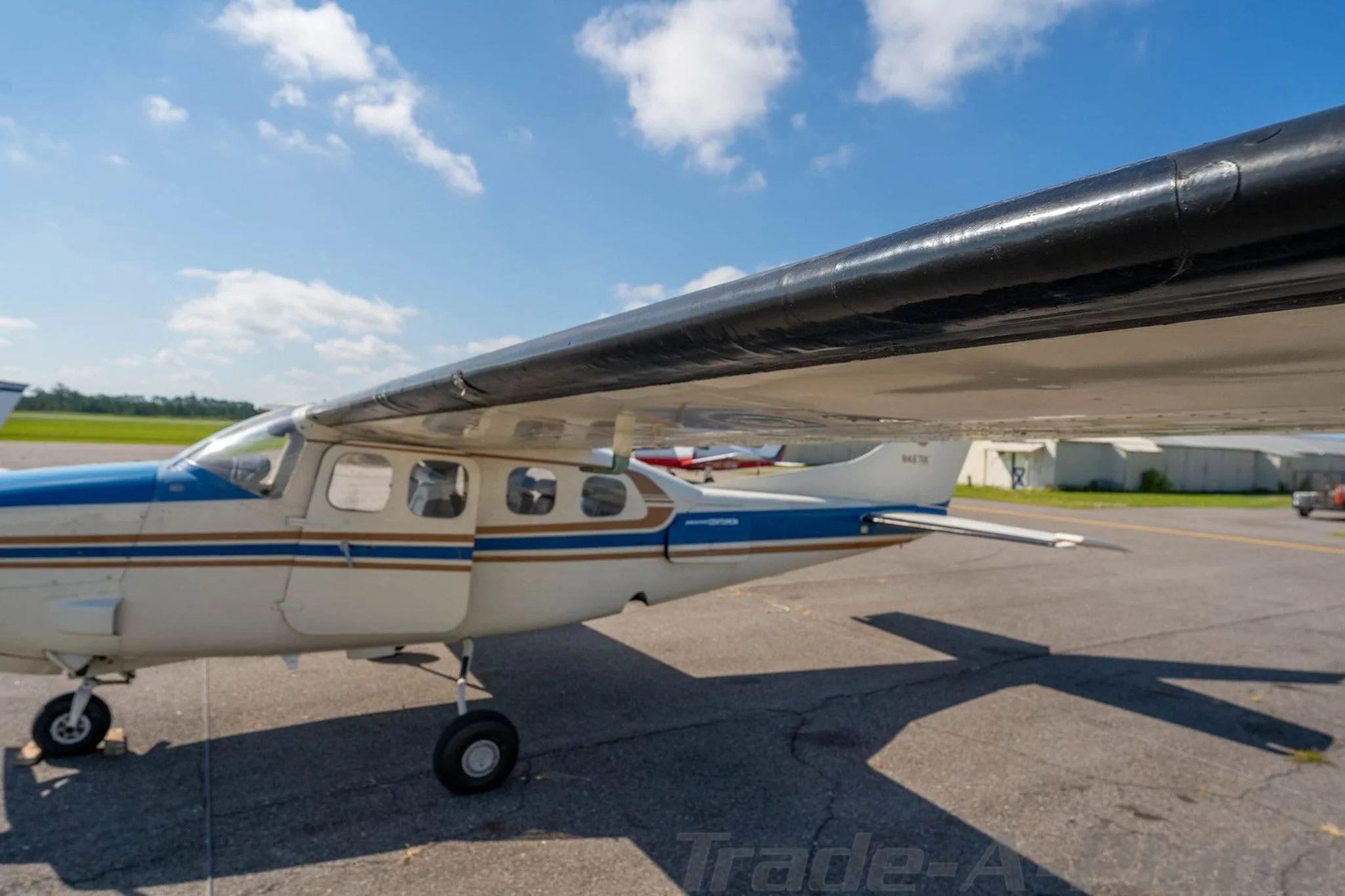 A side view of the white and blue Cessna Centurion N4671K aircraft on the tarmac, showing the open passenger door and wing.