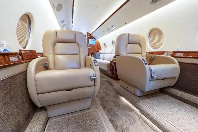 Two light-colored leather seats in the forward cabin of a private jet, with a view towards the rear seating area.