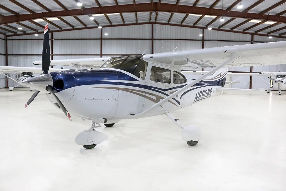 A white and blue Cessna Skylane N890MB is parked in a hangar, viewed from the front-left.