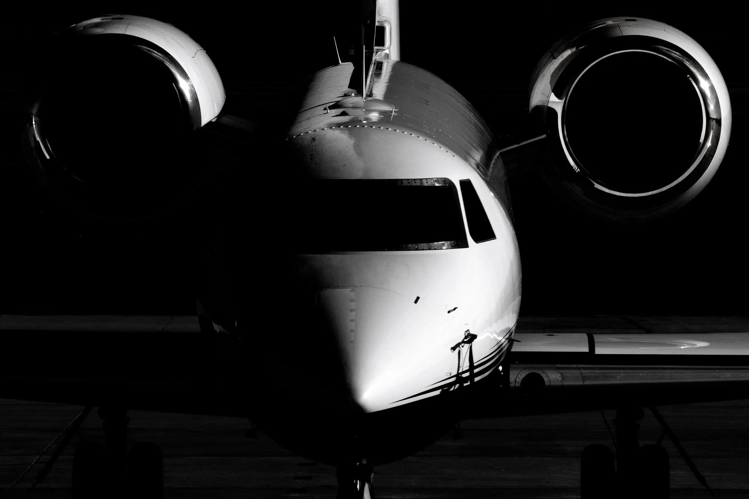 A black and white, head-on view of a business jet, highlighting its nose, cockpit, and engine nacelles against a dark background.