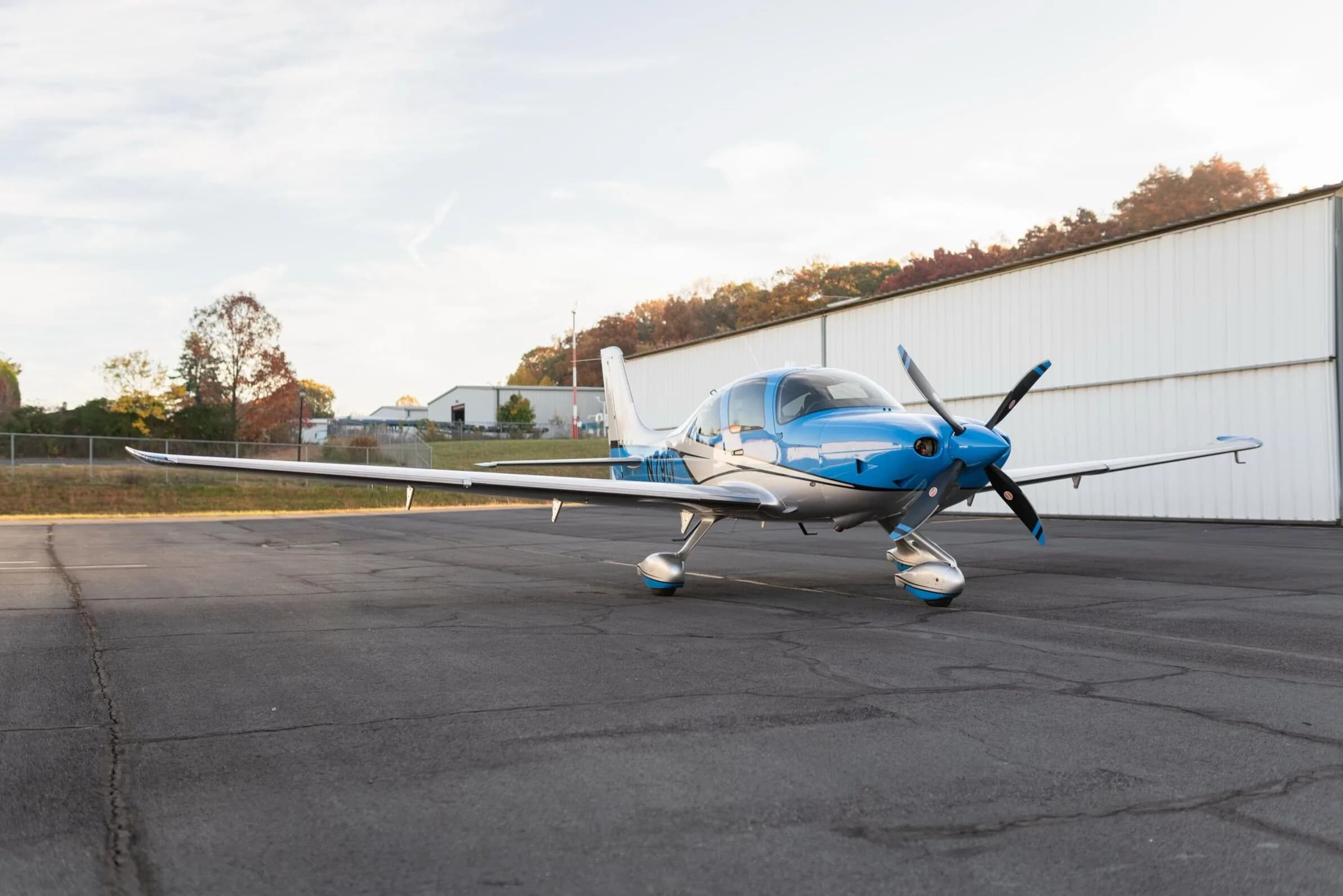 A blue and silver Cirrus SR22T GTS aircraft, registration N79CV, parked on an airfield with hangars and trees in the background.