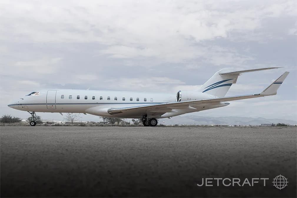 A white Bombardier Global 6000 business jet with blue and grey stripes on the tail, parked on an airfield under a cloudy sky.