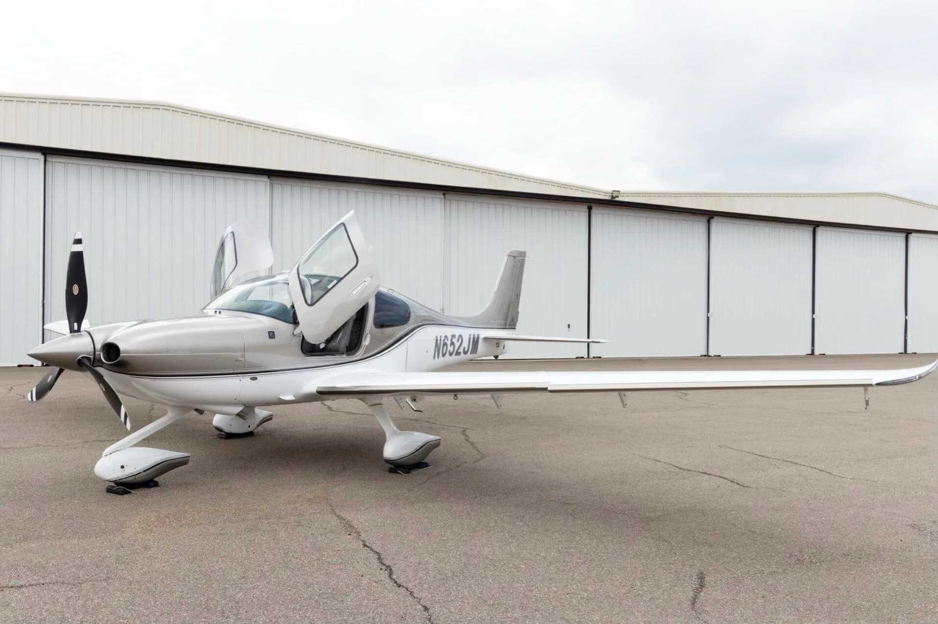 A three-quarter front view of a silver and white Cirrus SR22 aircraft with N652JM registration and both gull-wing doors open.