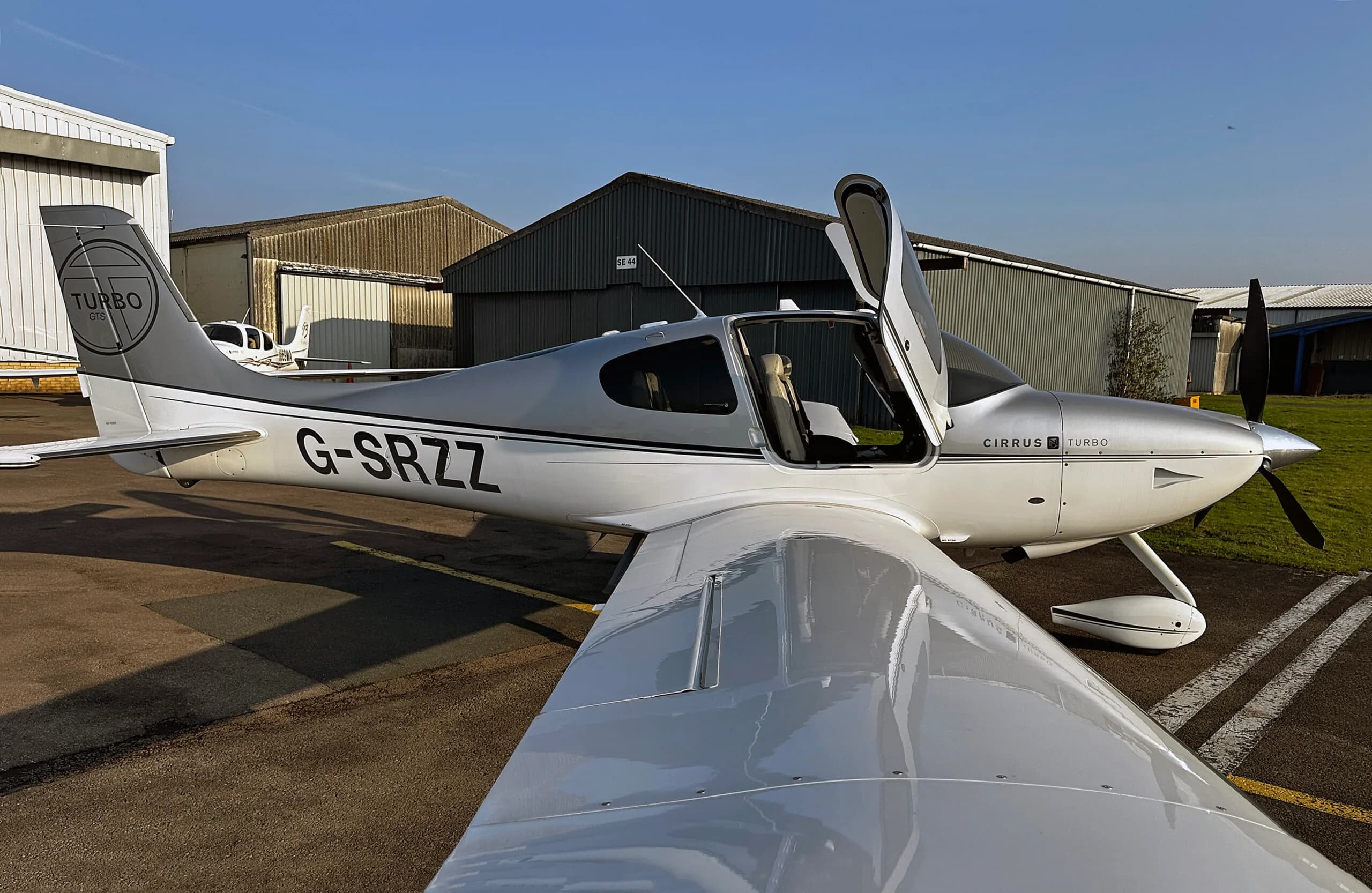A silver and white Cirrus SR22 Turbo aircraft with registration G-SRZZ and an open door, viewed from the wing.