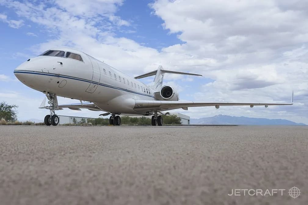 A low-angle front view of a white Bombardier Global 6000 business jet with blue and grey stripes, parked on an airfield.