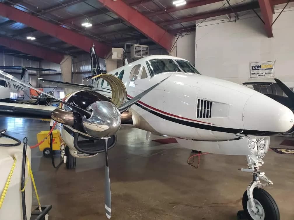 The front section of a white twin-engine turboprop aircraft with red and black stripes, parked inside a hangar.