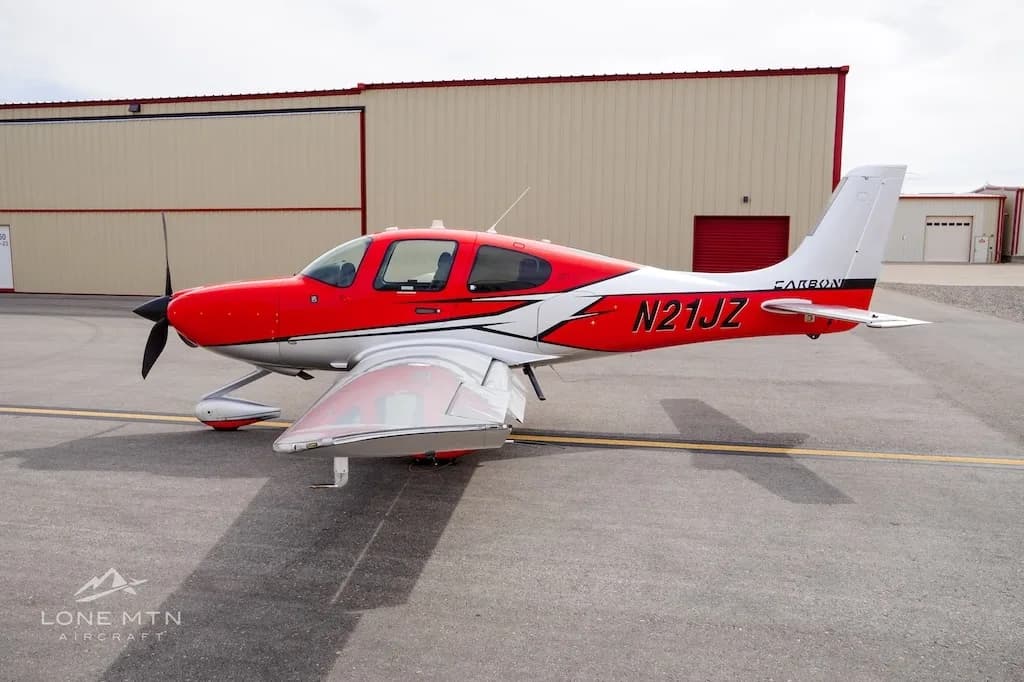 A side view of a red and silver Cirrus SR22 aircraft parked on an asphalt ramp in front of hangars.