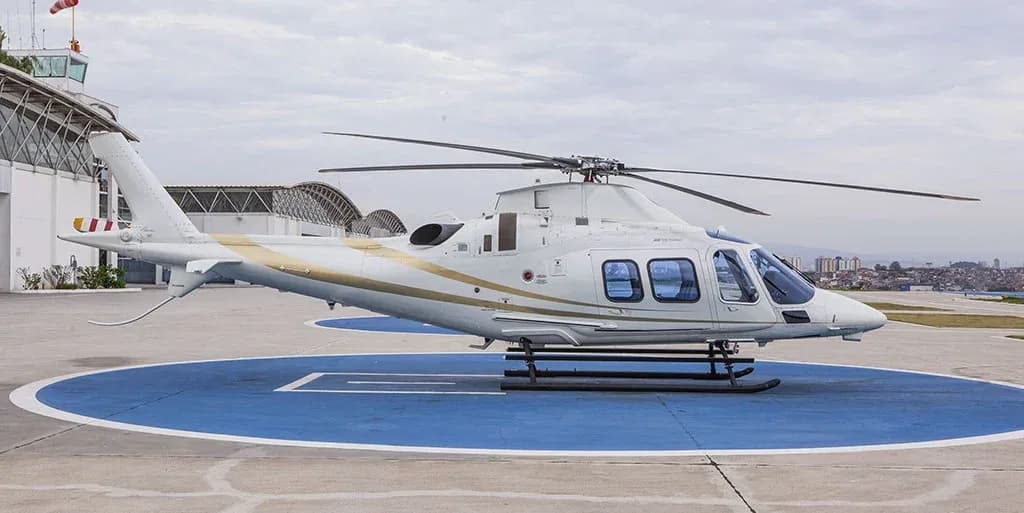 A white helicopter with gold stripes is parked on a blue helipad, viewed from the side with a control tower in the background.