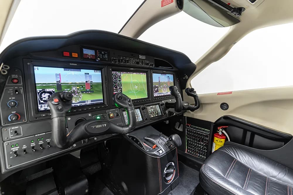 A side view of the TBM aircraft cockpit, showing the instrument panel, yokes, and a fire extinguisher.