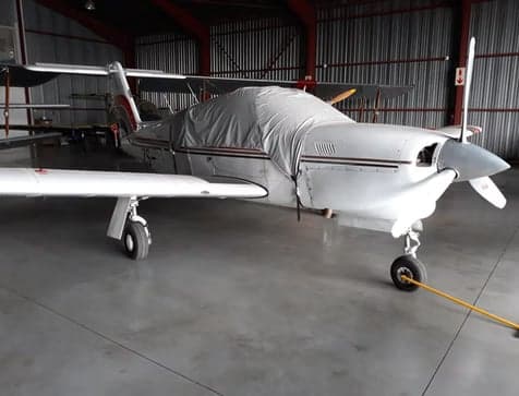 A white single-engine propeller aircraft, partially covered, parked inside a hangar with another aircraft in the background.