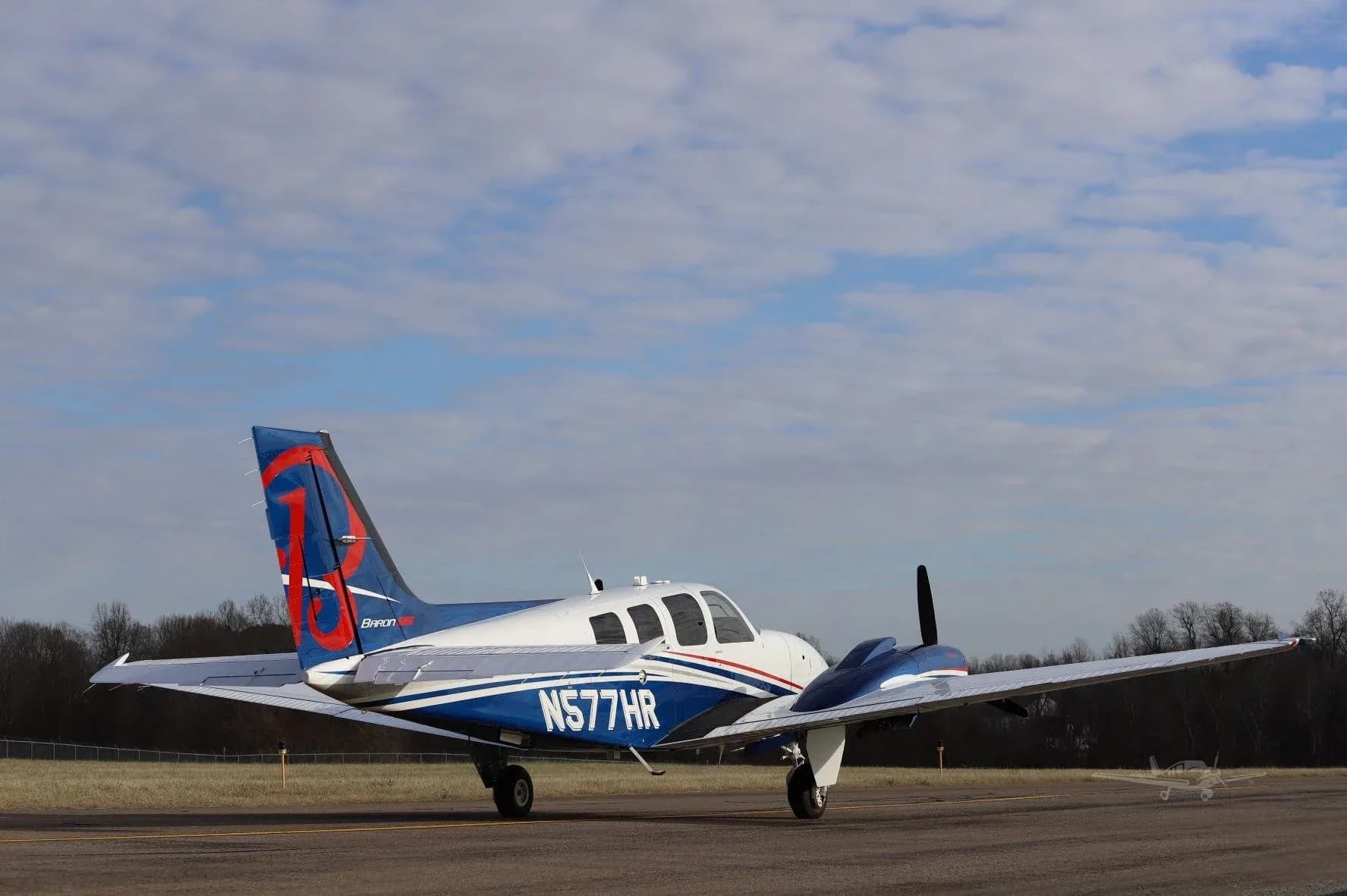 A white and blue Beechcraft Baron G58 (N577HR) with red tail accents, seen from the rear-right on a tarmac under a partly cloudy sky.
