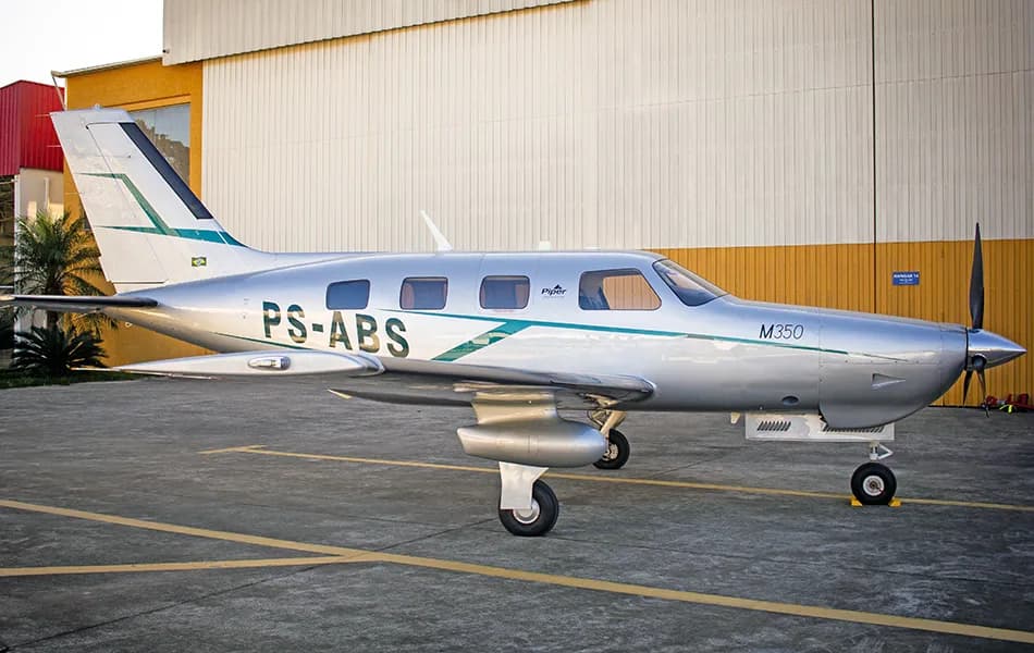 A silver Piper M350 aircraft with green and dark blue stripes is parked on a tarmac in front of a hangar.