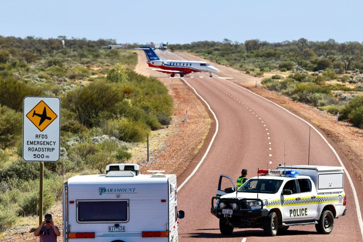 A Pilatus PC-24 aircraft parked on a remote runway with a police vehicle and caravan nearby.