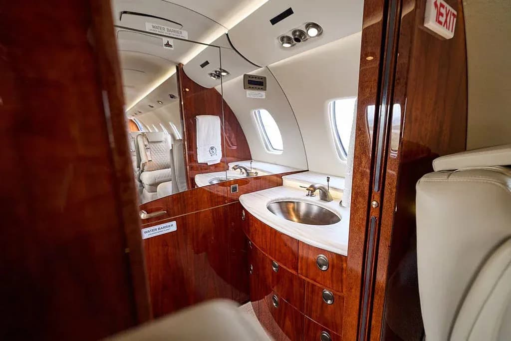 The aircraft lavatory features a polished wood vanity with a silver sink and a large mirror.