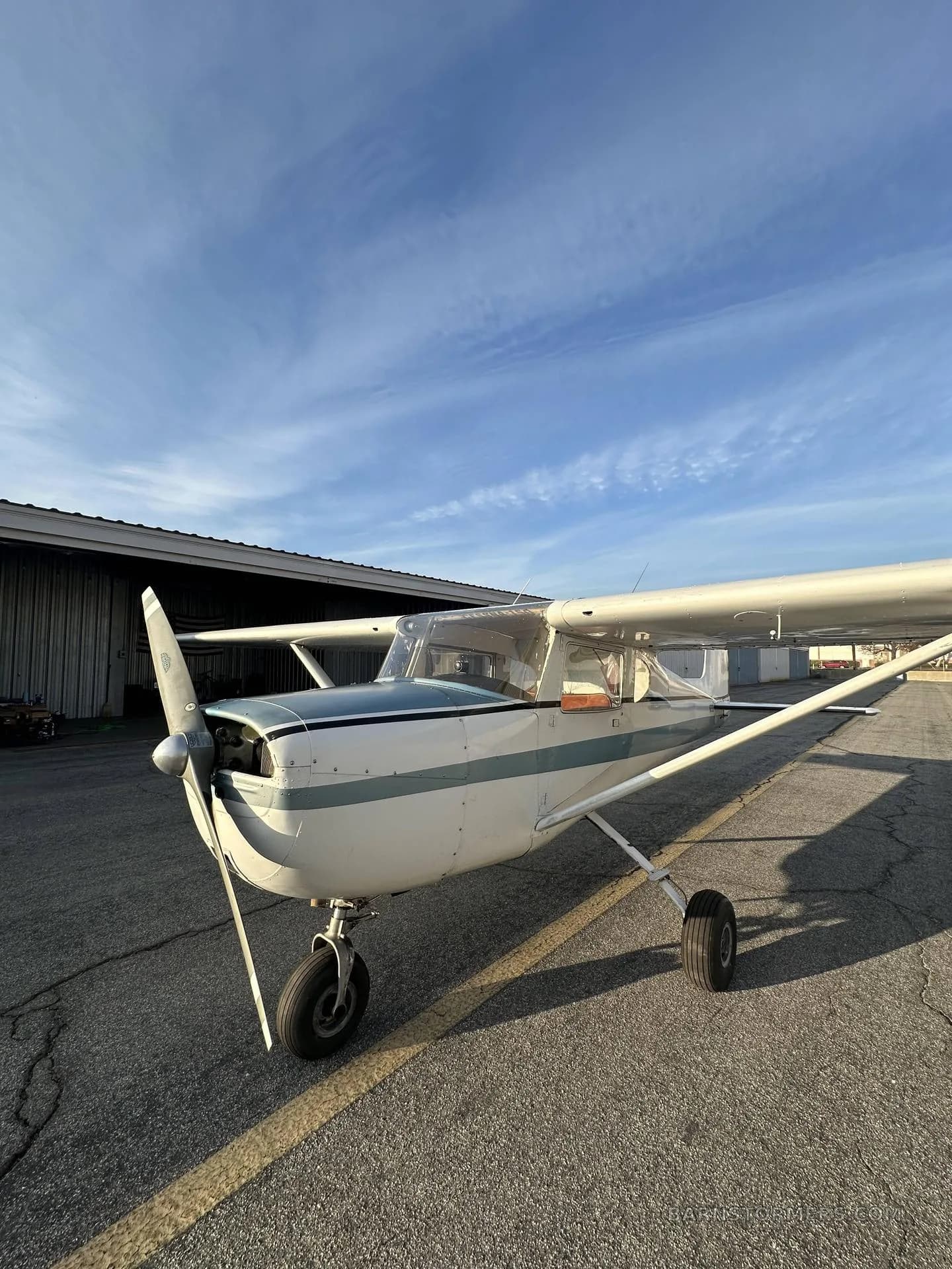 A white and blue Cessna 150/152 aircraft is parked on a tarmac next to a hangar under a blue sky.