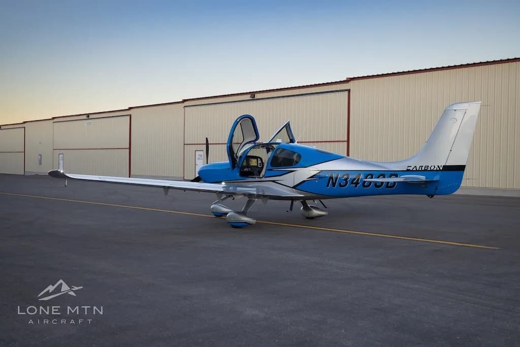 A three-quarter rear view of a blue and silver Cirrus SR22 aircraft with open doors parked on an airfield next to hangars.