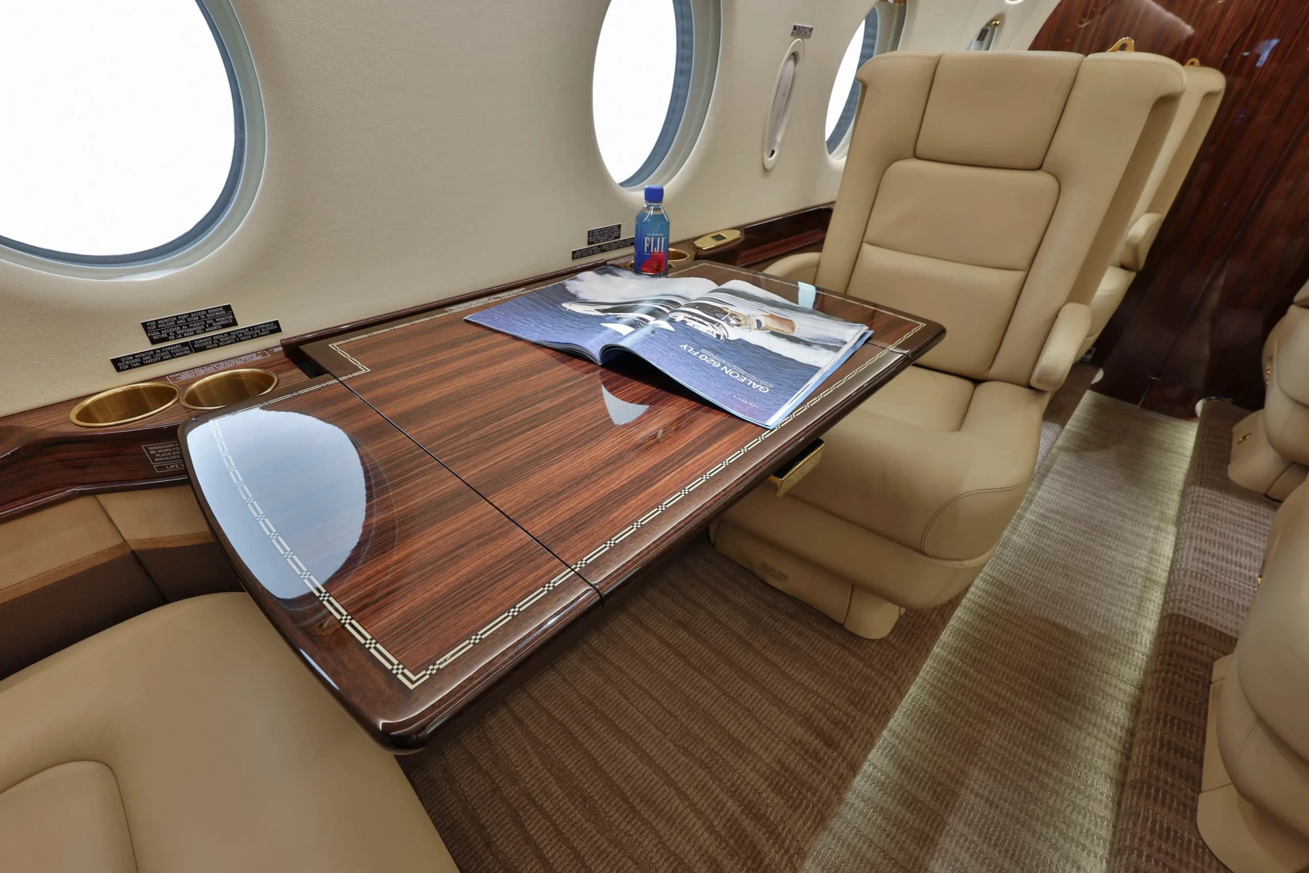 A luxurious aircraft cabin interior featuring a polished wooden table, beige leather seats, and a Fiji water bottle.