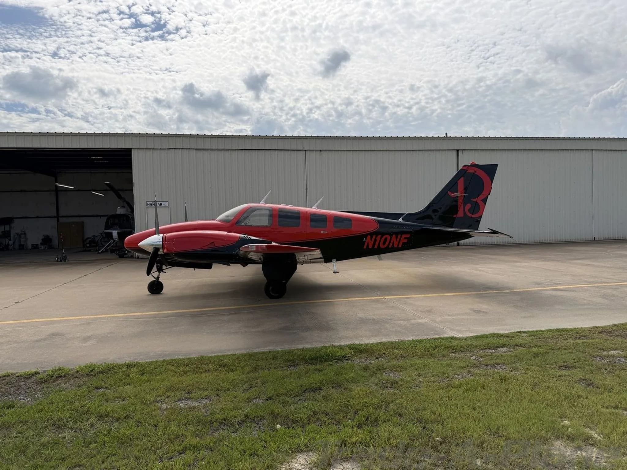 Side view of the red and black twin-engine propeller aircraft, N10NF, with a large red "13" on its tail, parked in front of a hangar.