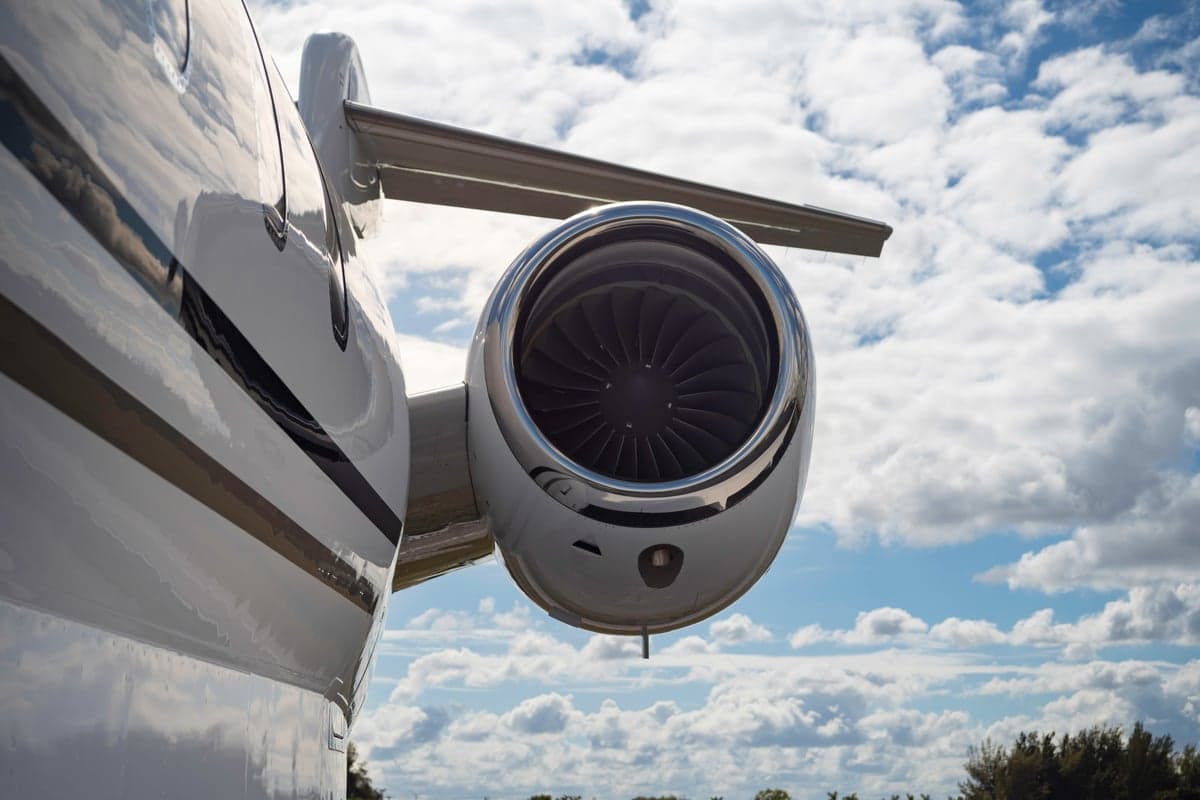A close-up of the rear-mounted engine of a white business jet against a blue sky with white clouds.