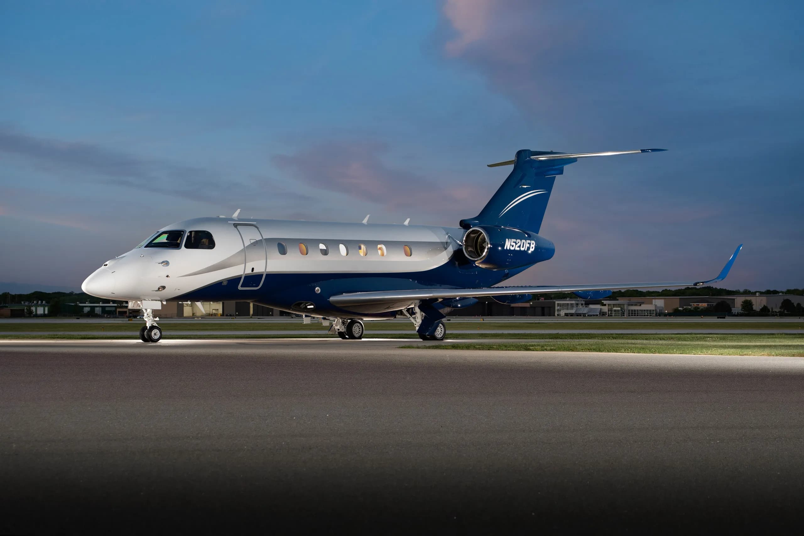 A silver and blue Embraer Phenom 300 business jet, registration N520FB, is parked on a tarmac at dusk with landing lights on.