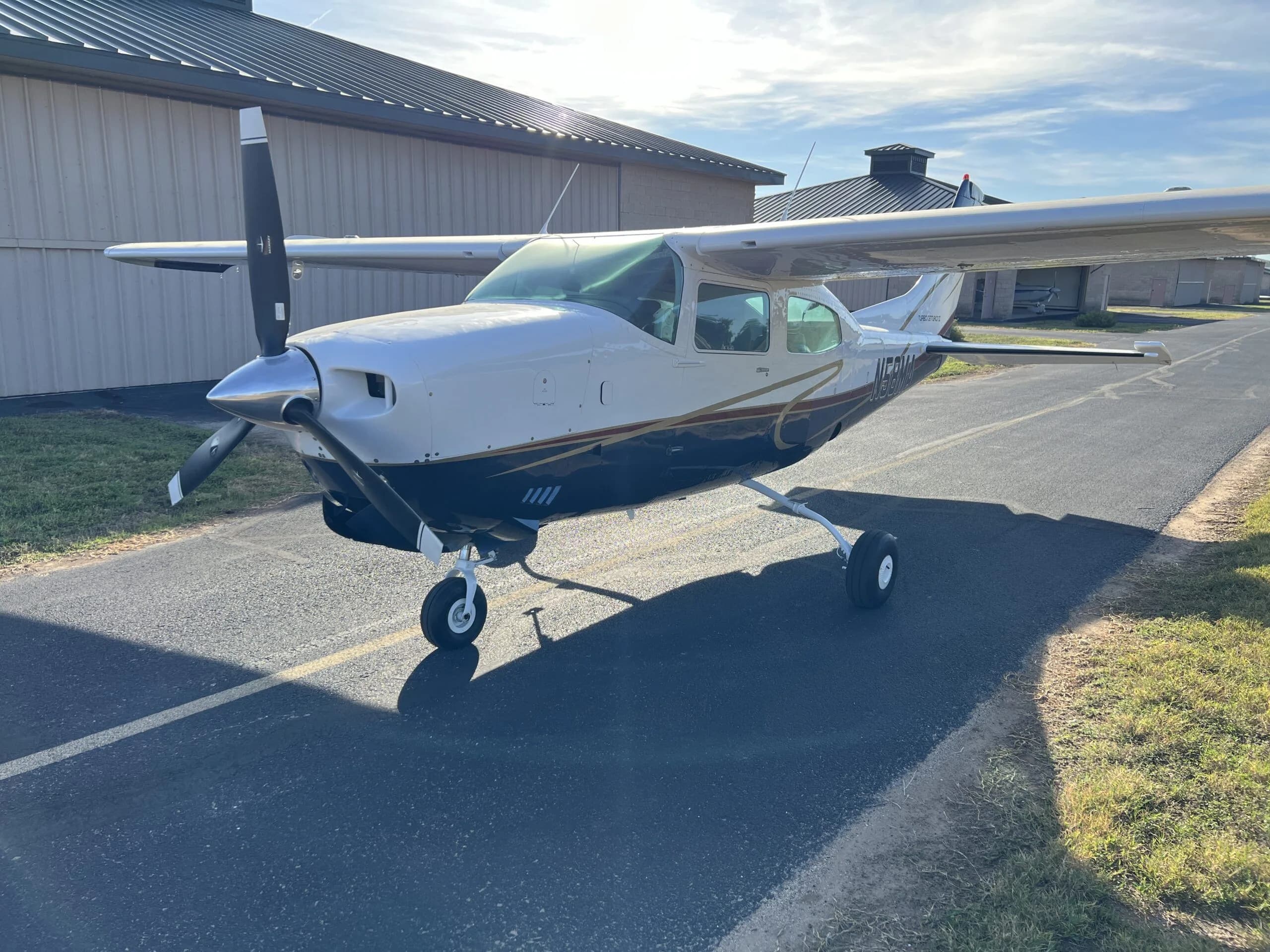 A white and blue Cessna 210, N58MA, with gold trim, parked on an asphalt taxiway next to a hangar.