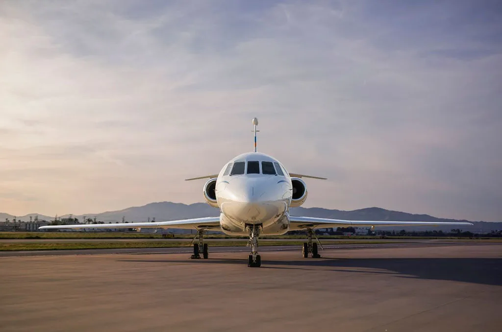 A white private jet with a clean paint scheme, viewed head-on on an airfield with mountains in the background.