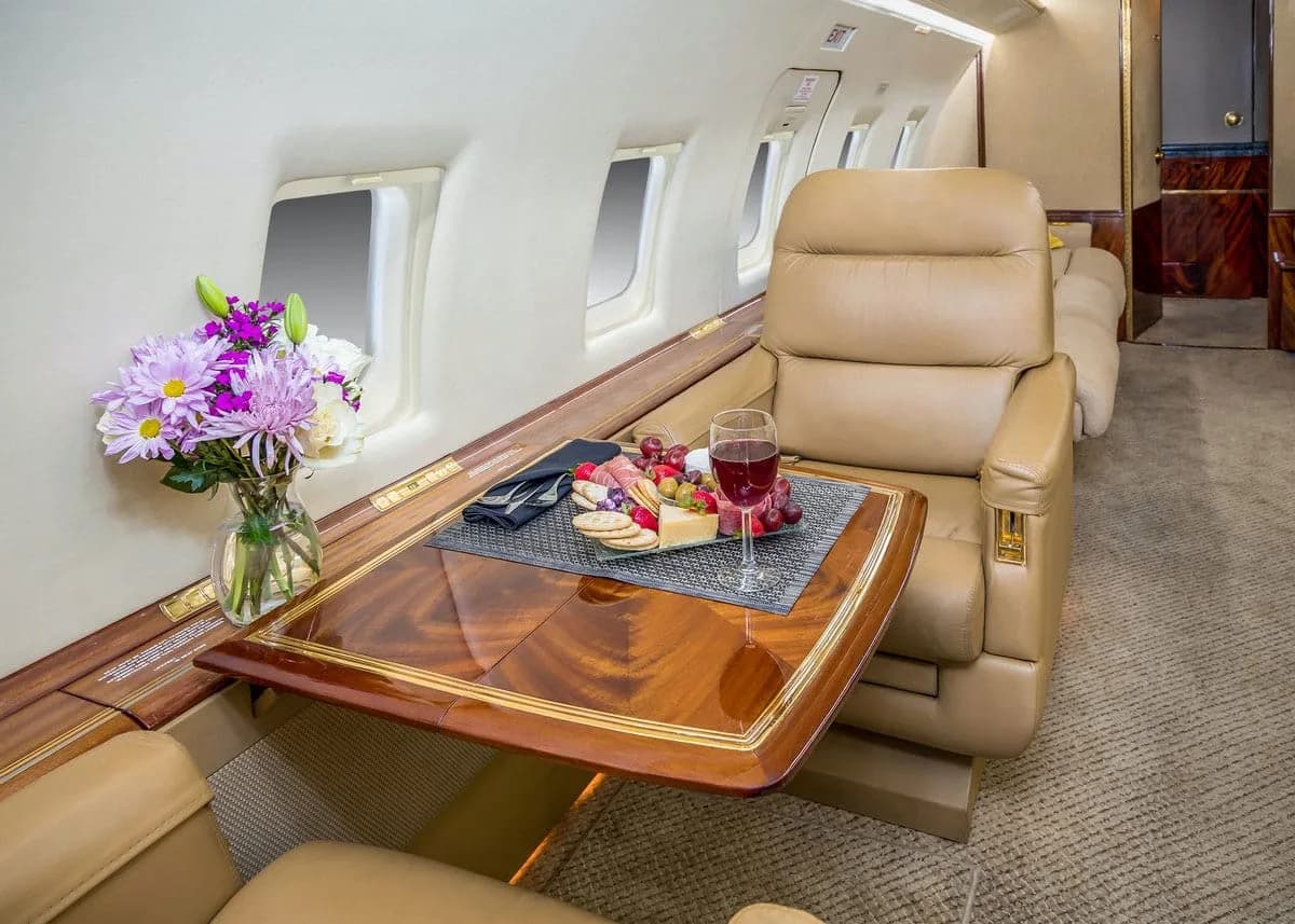 A comfortable tan leather seat next to a polished wooden table set with a charcuterie board, wine glass, and a vase of flowers inside an aircraft cabin.