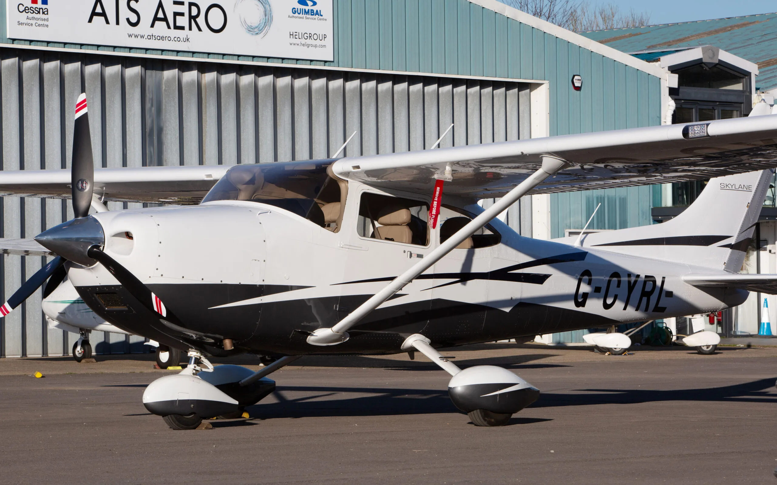 Side view of a white and black Cessna 182T (G-CYRL) parked on an airfield next to a hangar with "ATS AERO" signage.