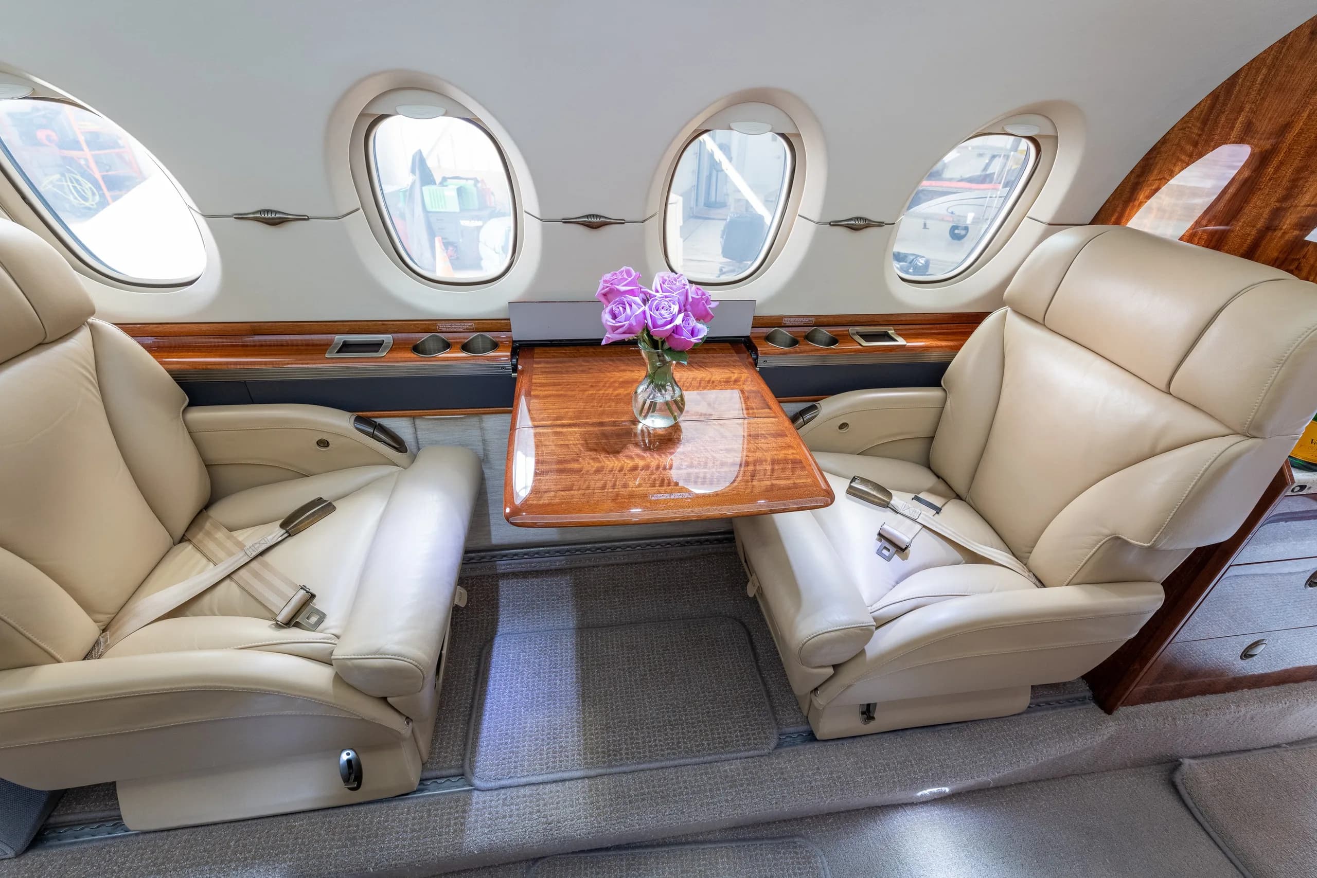 Two beige leather passenger seats facing each other with a wooden table and a vase of purple flowers in between.