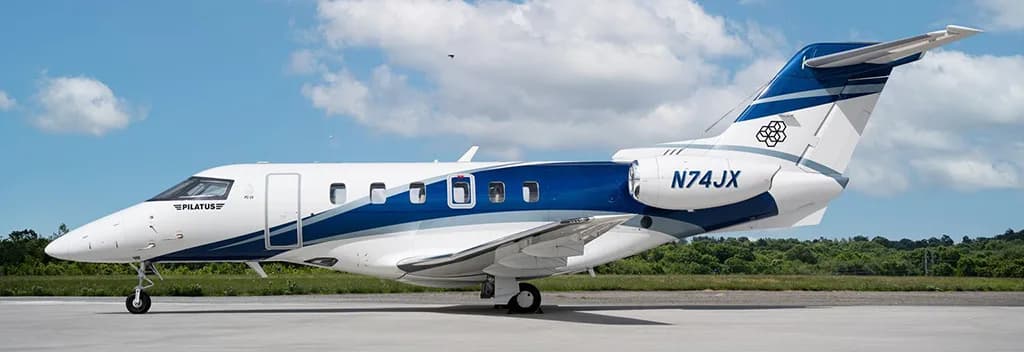 A Pilatus PC-24 business jet, registration N74JX, parked on an airfield with green trees in the background.