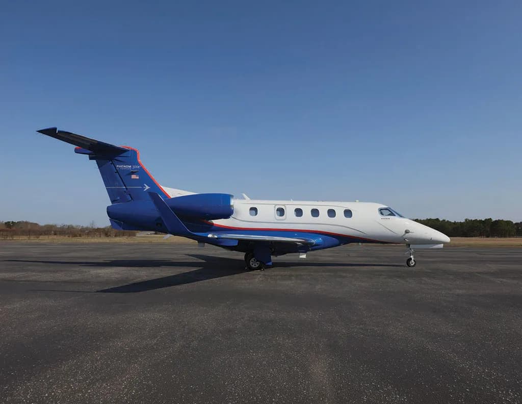 A white and blue Embraer Phenom 300 business jet is parked on an asphalt runway under a clear blue sky, viewed from the right side.